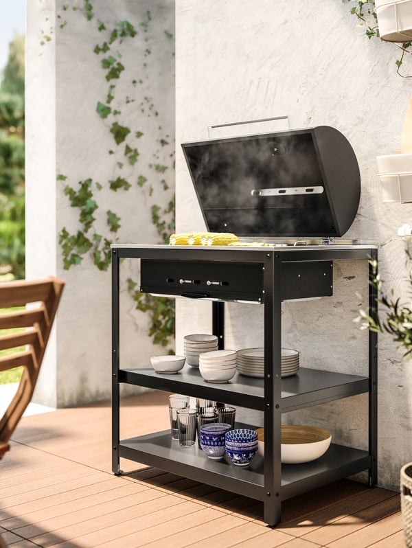 Parrilla GRILLSKÄR  apoyada en una pared blanca y junto a plantas.