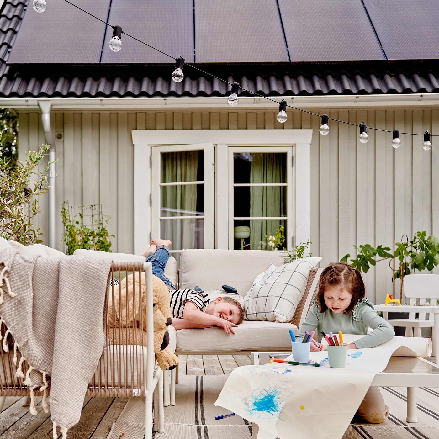 Zwei Kinder spielen draussen auf einer gemütlichen Terrasse, eines malt an einem Tisch, während das andere sich mit einem Teddybär auf einem Sofa entspannt.