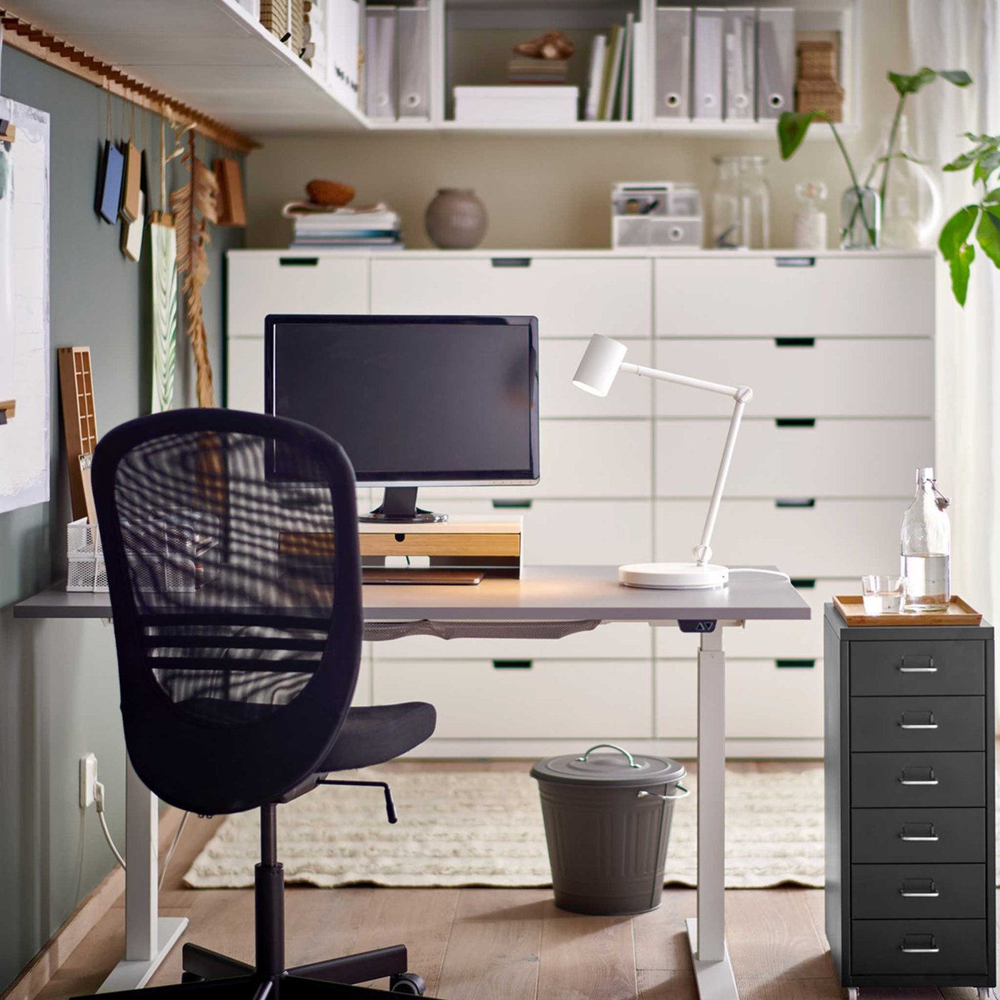 A black FLINTAN office chair and HELMER drawer unit by a grey RODULF sit/stand desk with a work lamp and computer screen.