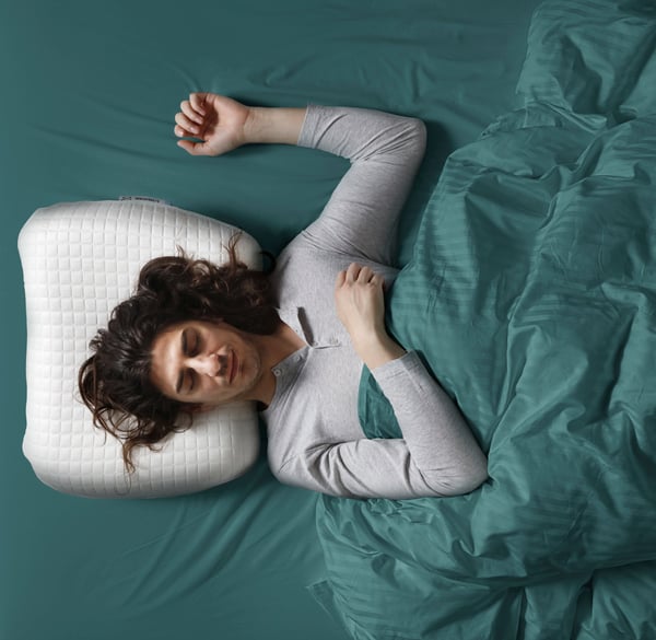 Vue en plongée sur une personne dormant sur le dos avec un bras levé sur le côté et la tête sur un oreiller ergonomique KLUBBSPORRE.