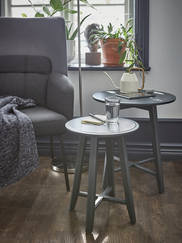 Dark blue-green nesting tables beside a gray armchair, with a glass of water, tablet and watering can on them.