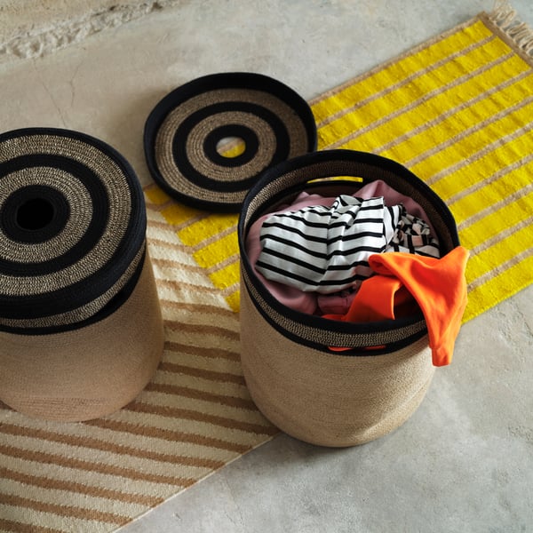 Two MÄVINN laundry baskets stand on a yellow rug. One has a lid on, the other is filled with laundry with the lid next to it.