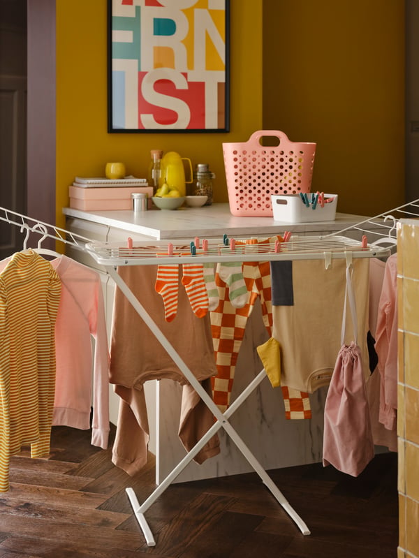A white MULIG drying rack with clothes held by clothes pegs and hangers, plus a pink flexible laundry basket on a worktop.