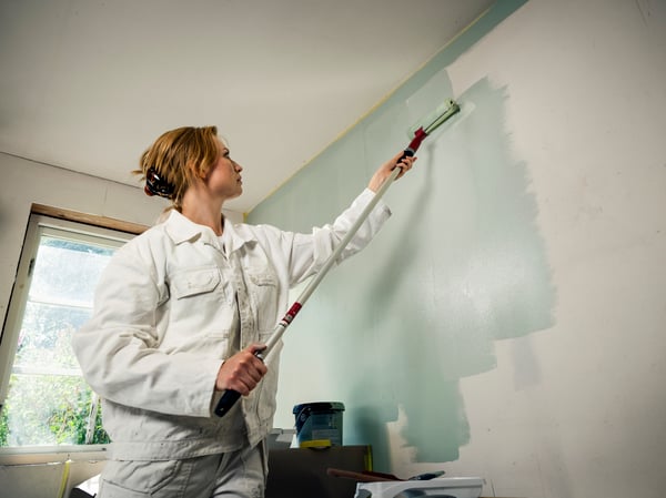 A woman in white painter's overalls uses a long-handled roller to paint a wall light blue.