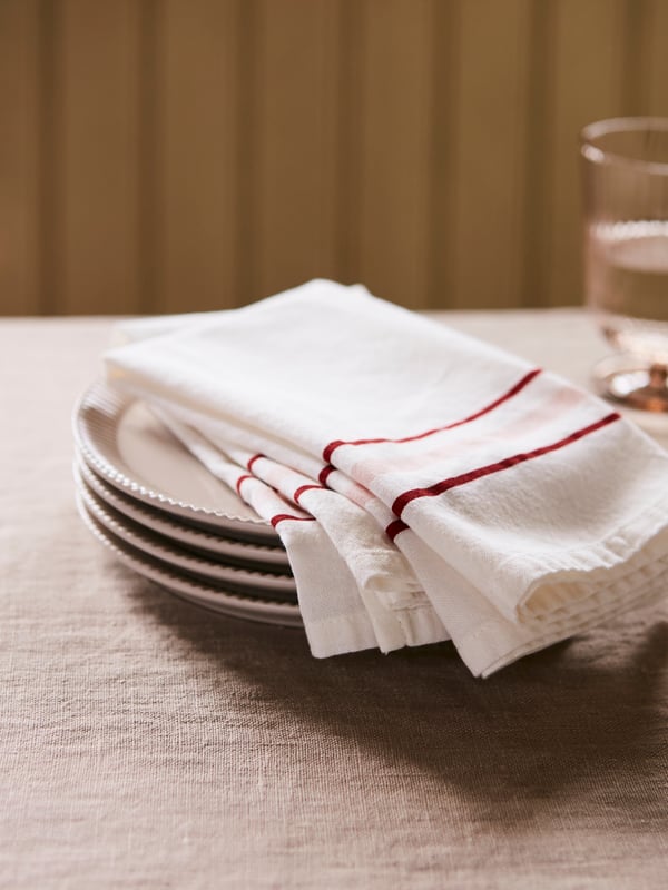 Two ANLEDNING textile napkins in white with red and pink stripes on the edge are lying on a stack of plates on a table.