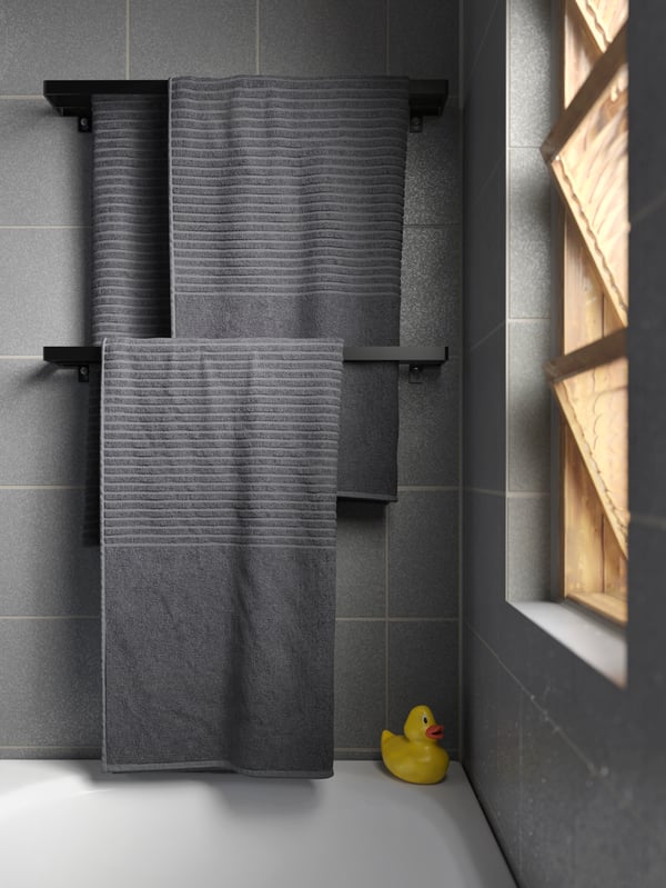 Grey towels hang on two black towel rails that are wall-mounted over a bathtub. A rubber duck stands in the bathtub’s corner.