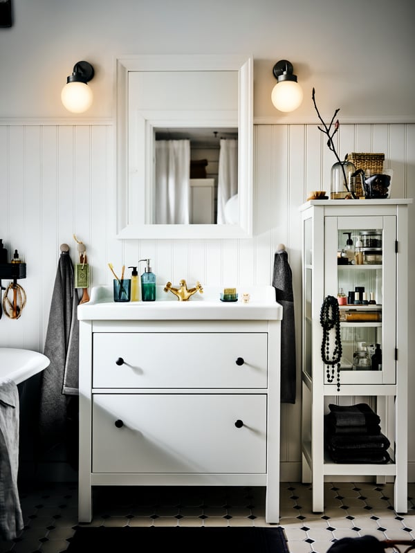 A HEMNES/RÄTTIVIKEN wash-stand with two drawers in a sunny white country-style bathroom with a mirror and wall lights above.
