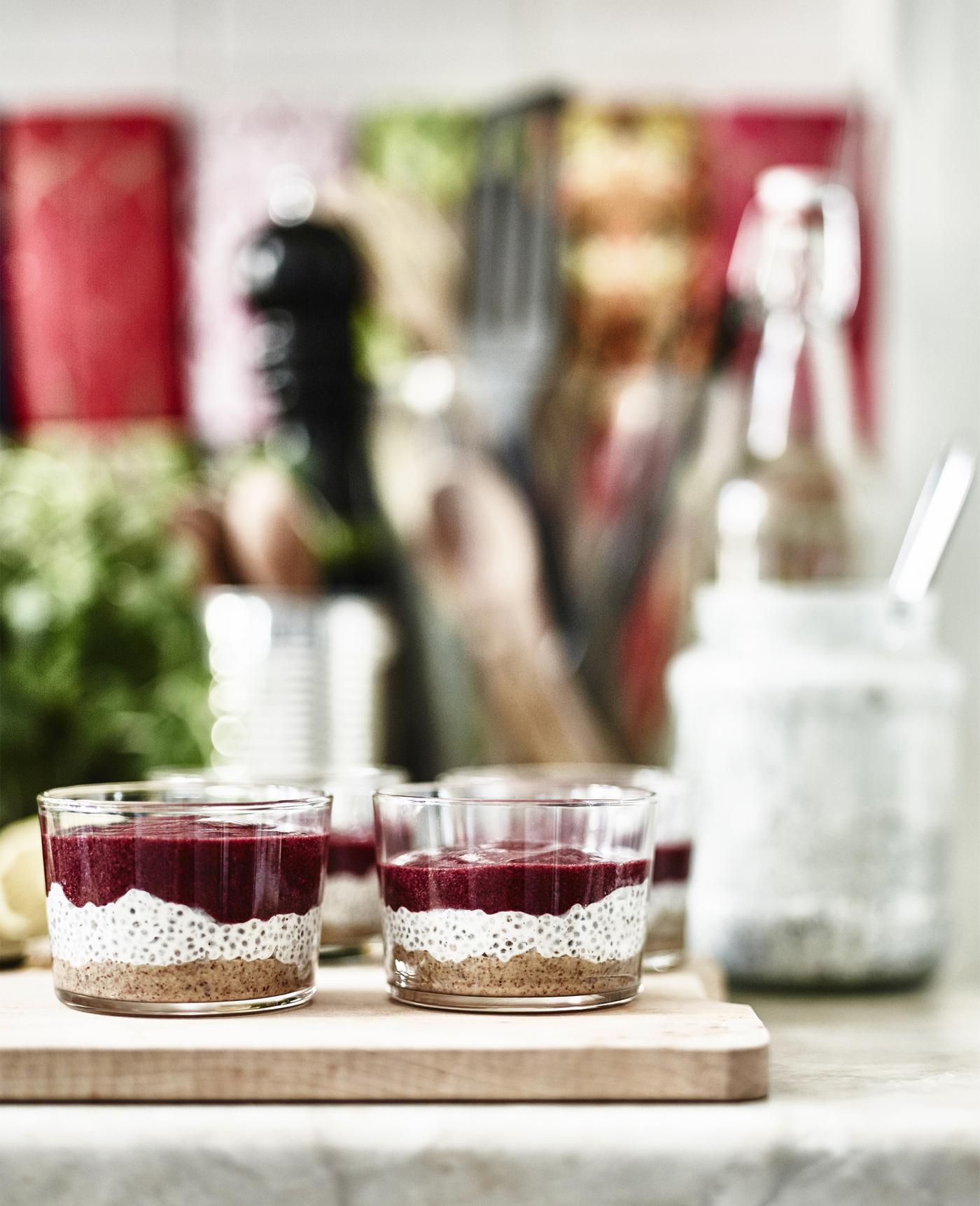 Zwei Gläser mit Schichtdessert aus Chiasamen, weisser Creme und rotem Fruchtpüree auf Holzbrett, unscharfer Hintergrund.