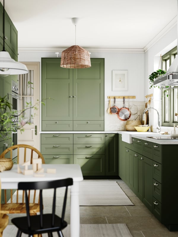 A bright kitchen with AXSTAD fronts in grey-green, white countertops, and a farmhouse sink. Natural light fills the space.