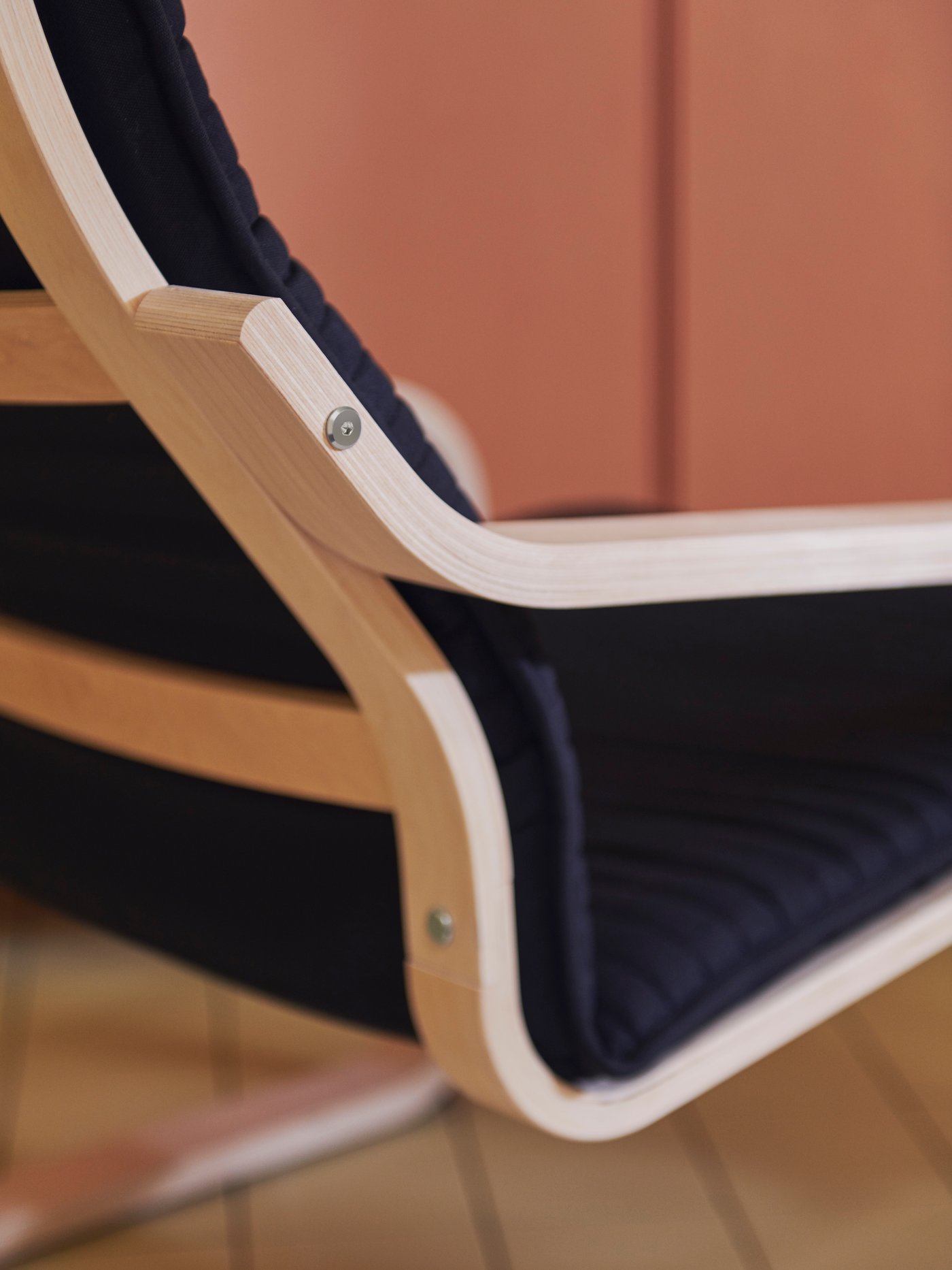 A close-up of the back and side of a POÄNG armchair showing its curved birch veneer frame and black cushion by a pink wall.