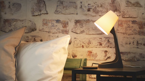 A dark grey/white SVALLET work lamp on a bedside table next to a bed in a bedroom.