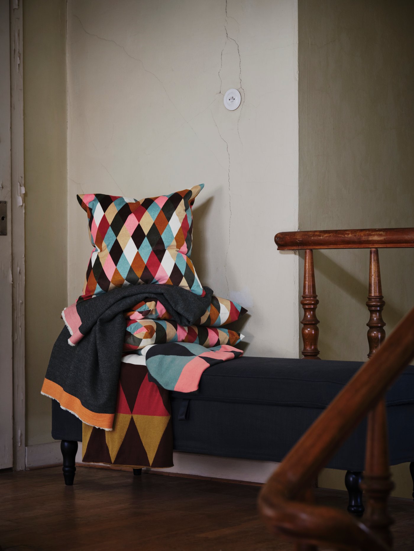 Diamond-patterned DEKORERA blankets and cushions in bright colours are piled on top of a bench by a set of stairs.