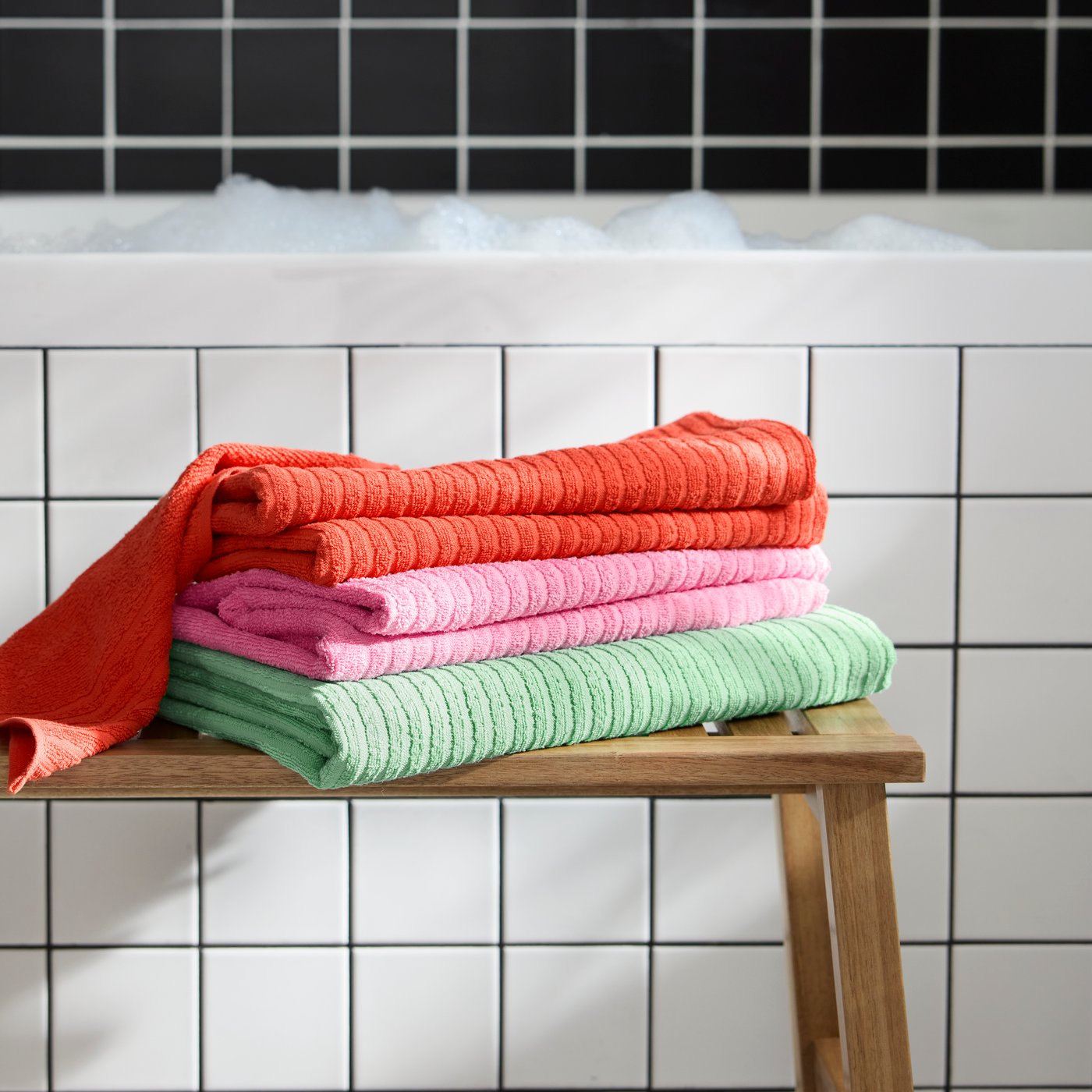 Three VÅGSJÖN bath towels in green, pink and red colours, folded on a wooden stool next to a white tiled bathtub.