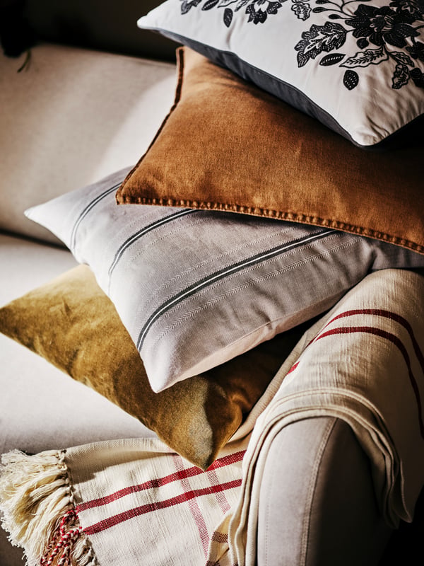 Quatre coussins dont un avec une housse de coussin STORTIMJAN reposant à l’extrémité d’un canapé 2 places LINANÄS beige, un plaid jeté sur l’accoudoir.