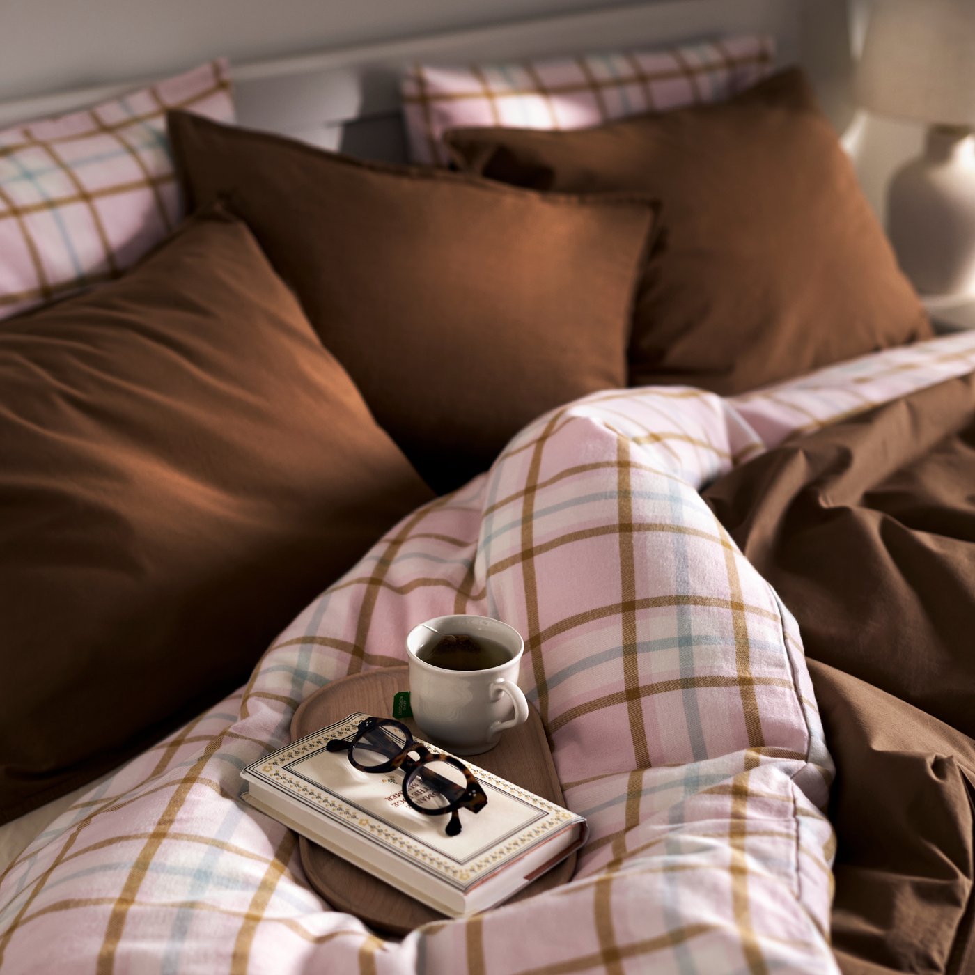A bed with pink flannel PEROVSKA duvet cover and brown SANELA cushions and a tray with a book, cup of coffee and glasses.