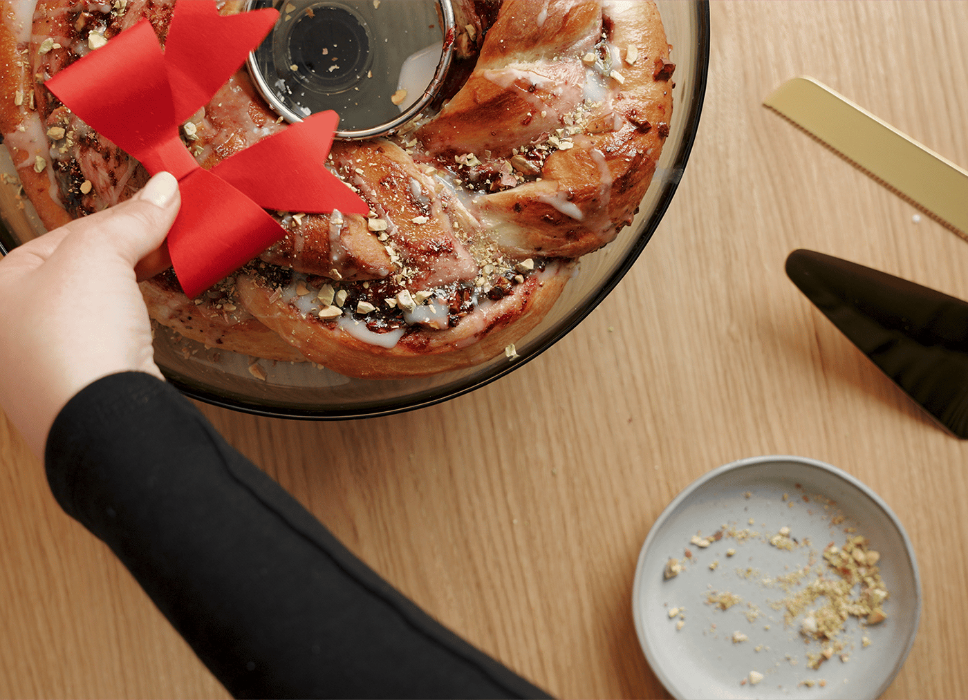 A hand places a red ribbon on a festive, glazed pastry wreath with nuts. Nearby, a golden cake server and a small plate with crumbs lie on a wooden table.