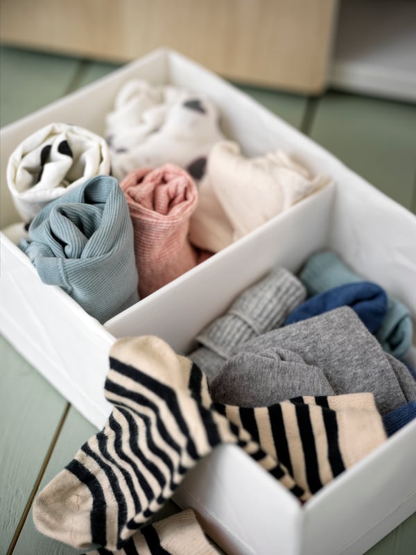 Une boîte de rangement STUK blanche à deux compartiments contenant des chaussettes et vêtements multicolores.