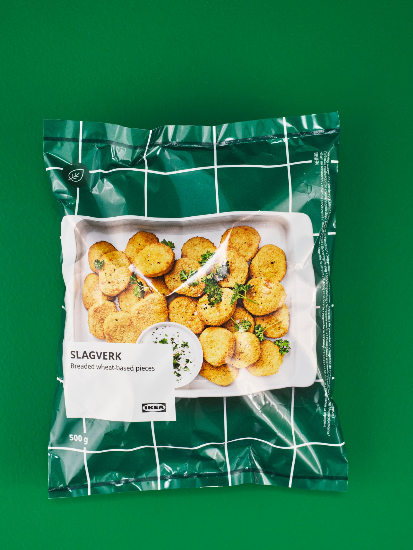 Un aliment emballé sur un arrière-plan vert, avec, derrière un volet transparent, des collations rondes à base de pommes de terre, garnies d’herbes, et étiqueté « SLAGVERK Smashed Herb-Infused Potatoes »