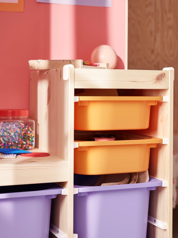 A light white stained pine TROFAST frame holds light orange and lilac boxes filled with art materials and toys.