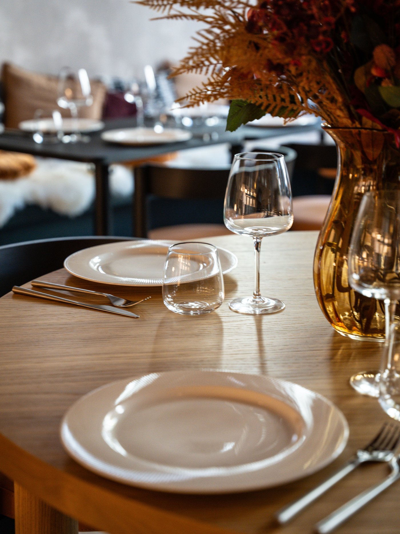 Gezellig gedekte houten tafel met wit servies, wijnglazen en een vaas met herfstbloemen in een warme restaurantsetting.