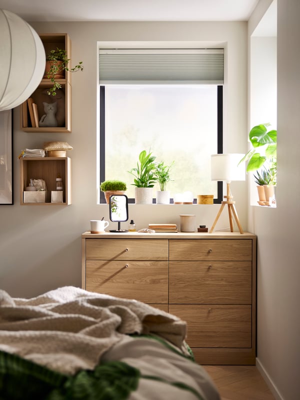 Vor einem Schlafzimmerfenster steht eine LAUTERS Tischleuchte auf einer TONSTAD Kommode mit 6 Schubladen. Am Fenster ist eine TREDANSEN Faltjalousie, verdunkelnd befestigt.