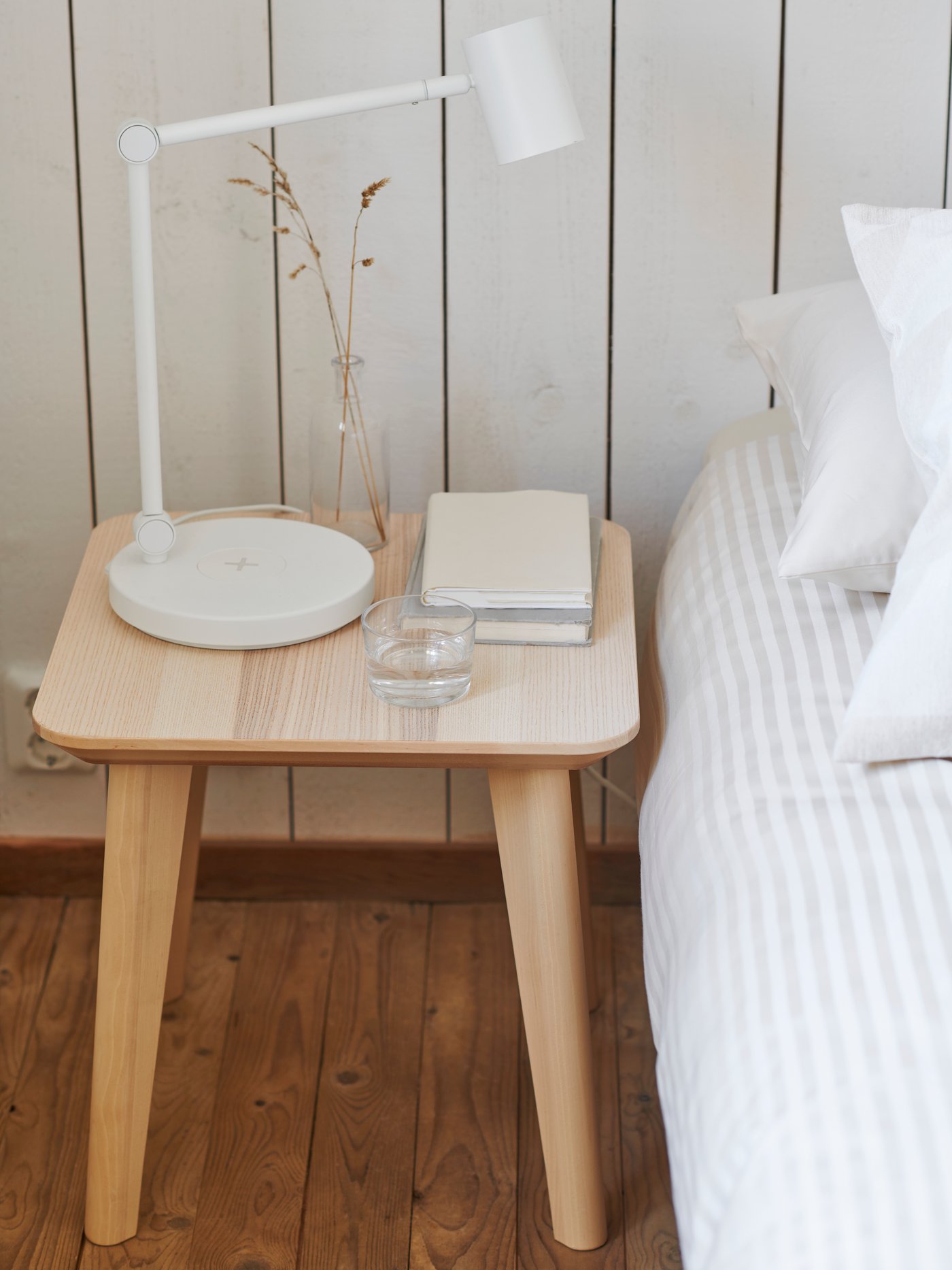 In einem Schlafzimmer im ländlichen Stil steht ein LISABO Beistelltisch mit einer NYMÅNE Arbeitsleuchte, einem Glas Wasser und Büchern neben einem Bett.