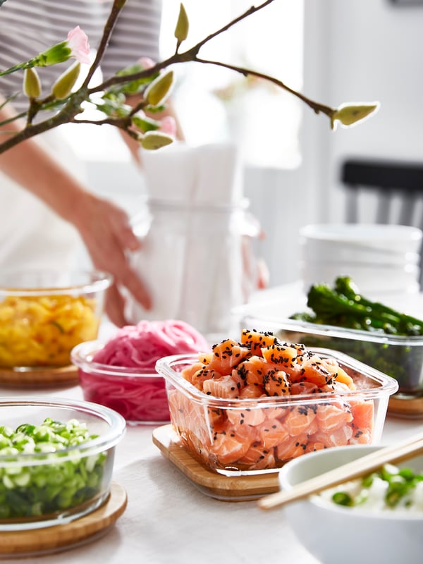 Plusieurs contenants en verre IKEA 365+, remplis de saumon, de différents légumes et de fruits, sont posés sur leurs couvercles en bambou.