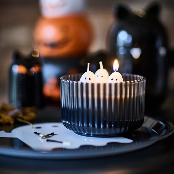 Et KUSTFYR duftlys i glas formet som tre spøgelser står på en KUSTFYR bakke med spøgelsesmotiv.