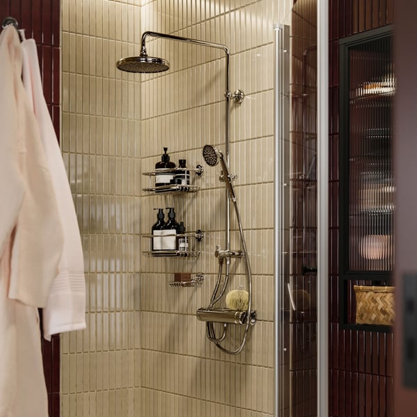 Two chrome-effect, traditional-looking BROFJÄRDEN shower shelves hold bottles in a beige-tiled shower with chrome fixtures.