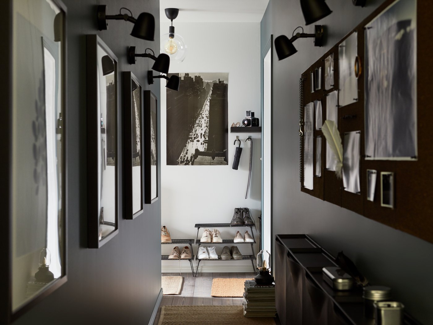 A narrow, modern hallway with dark gray walls, featuring a gallery of framed art on one side lit by black spotlights, and a large corkboard over a console table on the other.