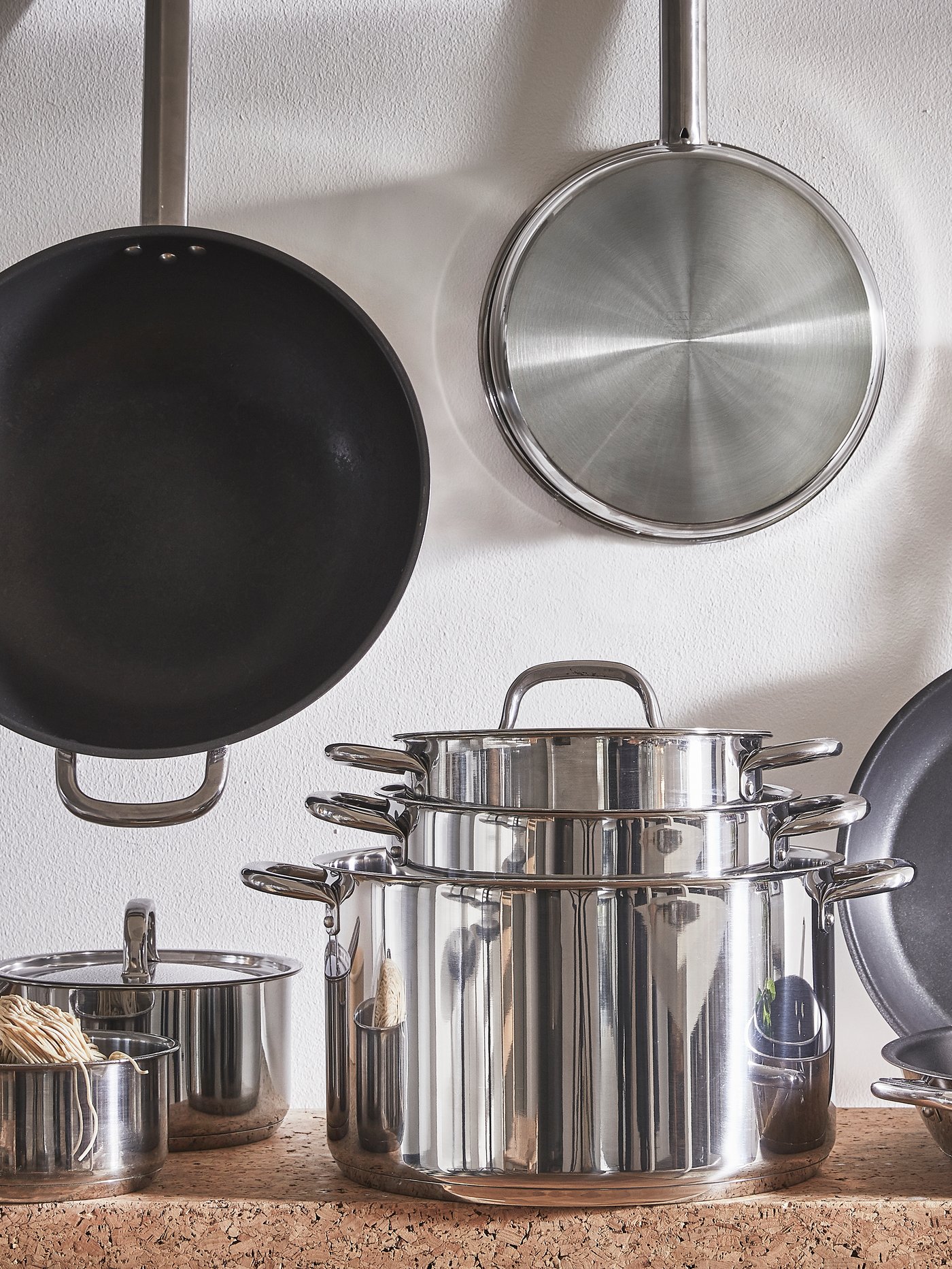 IKEA 365+ set posuđa za kuvanje iz 9 delova od nerđajućeg čelika na radnoj ploči s IKEA 365+ vok-tiganjem i tiganjem okačenim na zid u pozadini.