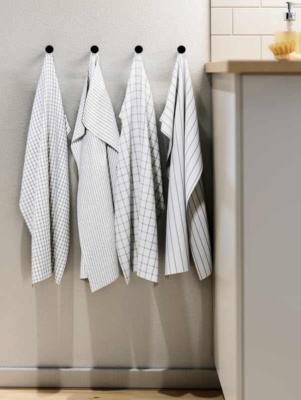 Four patterned, white-and-dark-grey RINNIG tea towels hang side-by-side on individual knobs by the end of a kitchen worktop.