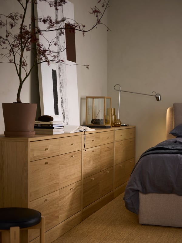 Drie TONSTAD ladekasten met 6 lades staan tegen een beige wand. Erop staan een MODERMOLN lamp, een plant en verschillende andere voorwerpen.