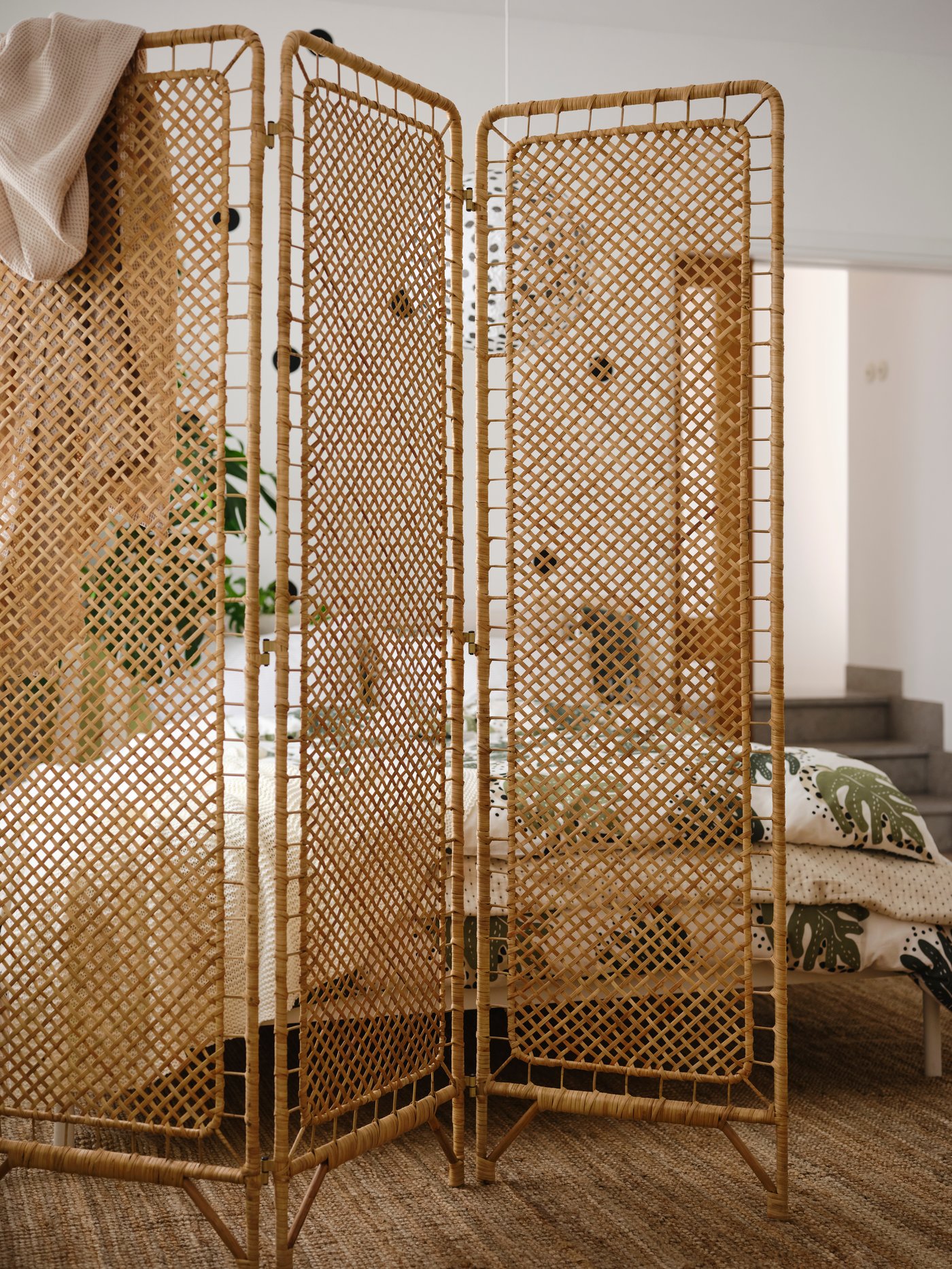 Room divider in braided rattan with several panels, placed in a bright room with wood and natural materials, where a bed and textiles are partially screened off behind.