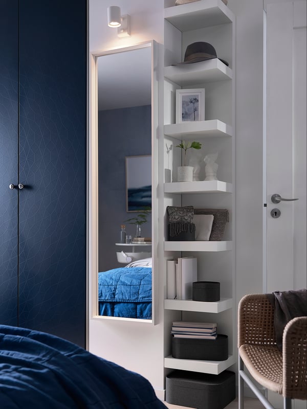 A vertically wall-mounted LACK wall shelf in a bedroom next to a mirror.
