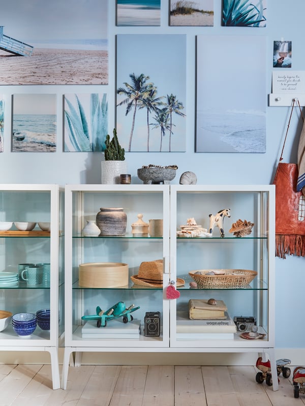 Two white glass door cabinets with decorative pieces inside, blue walls and beach prints hanging on the wall.