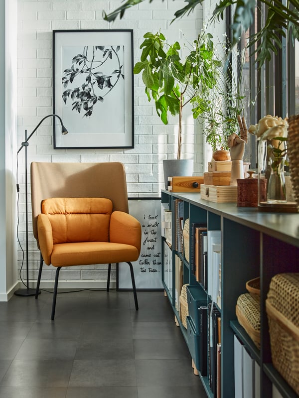 A reading nook with a yellow armchair, a black reading lamp, grey-turquoise cabinets by the window with books and boxes.