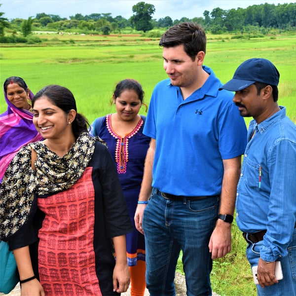Five people standing outside in a field. Two are wearing blue shirts and the other three are wearing brightly coloured saris.
