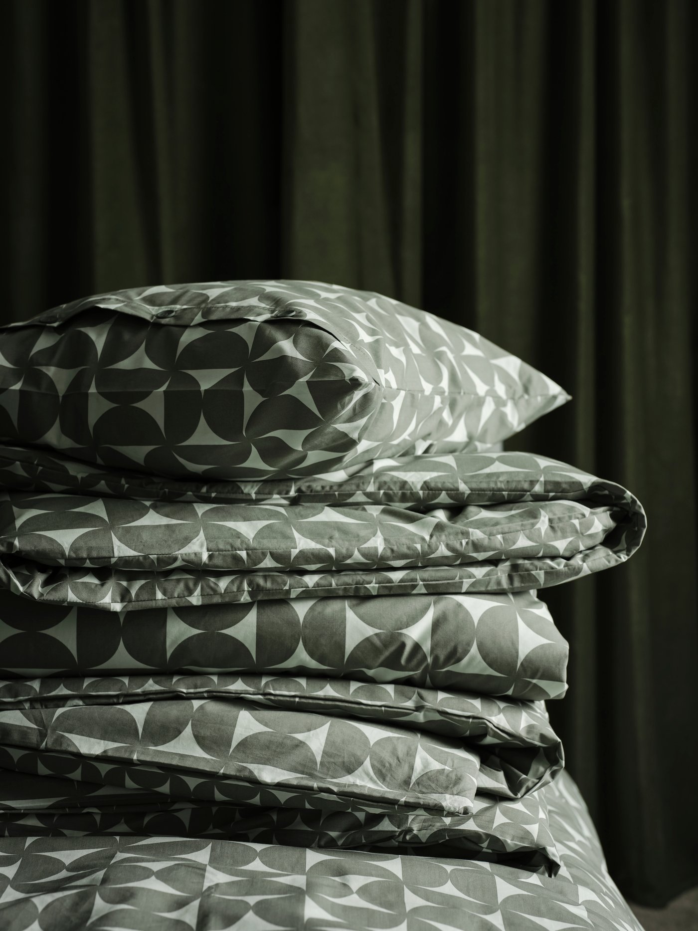 A folded duvet and a pillow covered in grey-green ÄNGSNEJLIKA bed linen in a pile on a bed in front of a SANELA curtain.