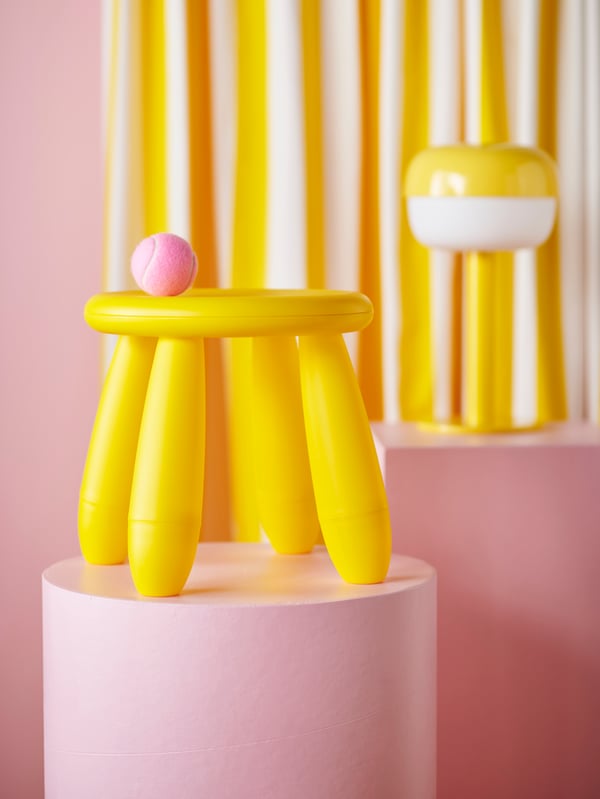 A yellow MAMMUT stool placed on a pink piedestal, and a yellow BLÅSVERK lamp in the back.