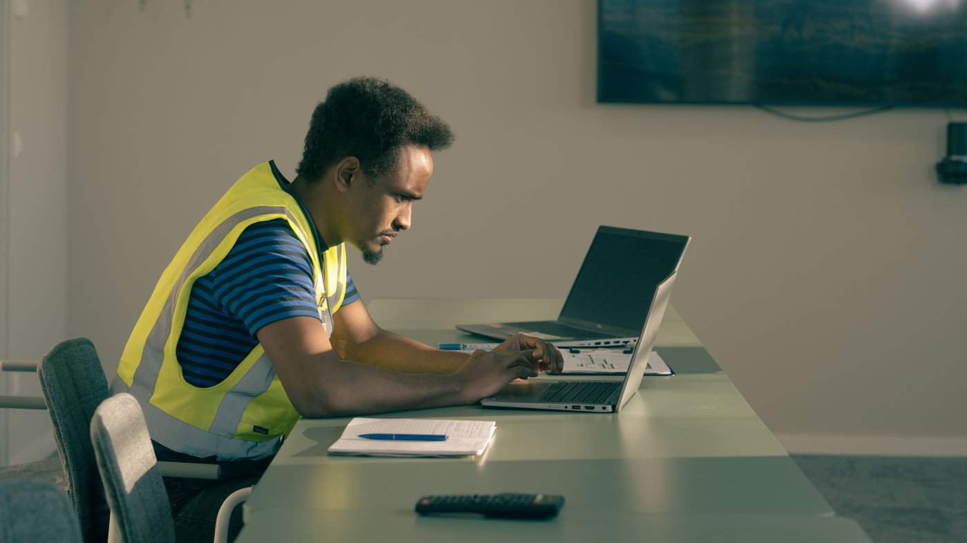 Abdi, der ein Hemd und eine Warnweste trägt, sitzt konzentriert an einem Konferenztisch an seinem Laptop. Daneben liegen ein Notizblock und eine Fernbedienung.
