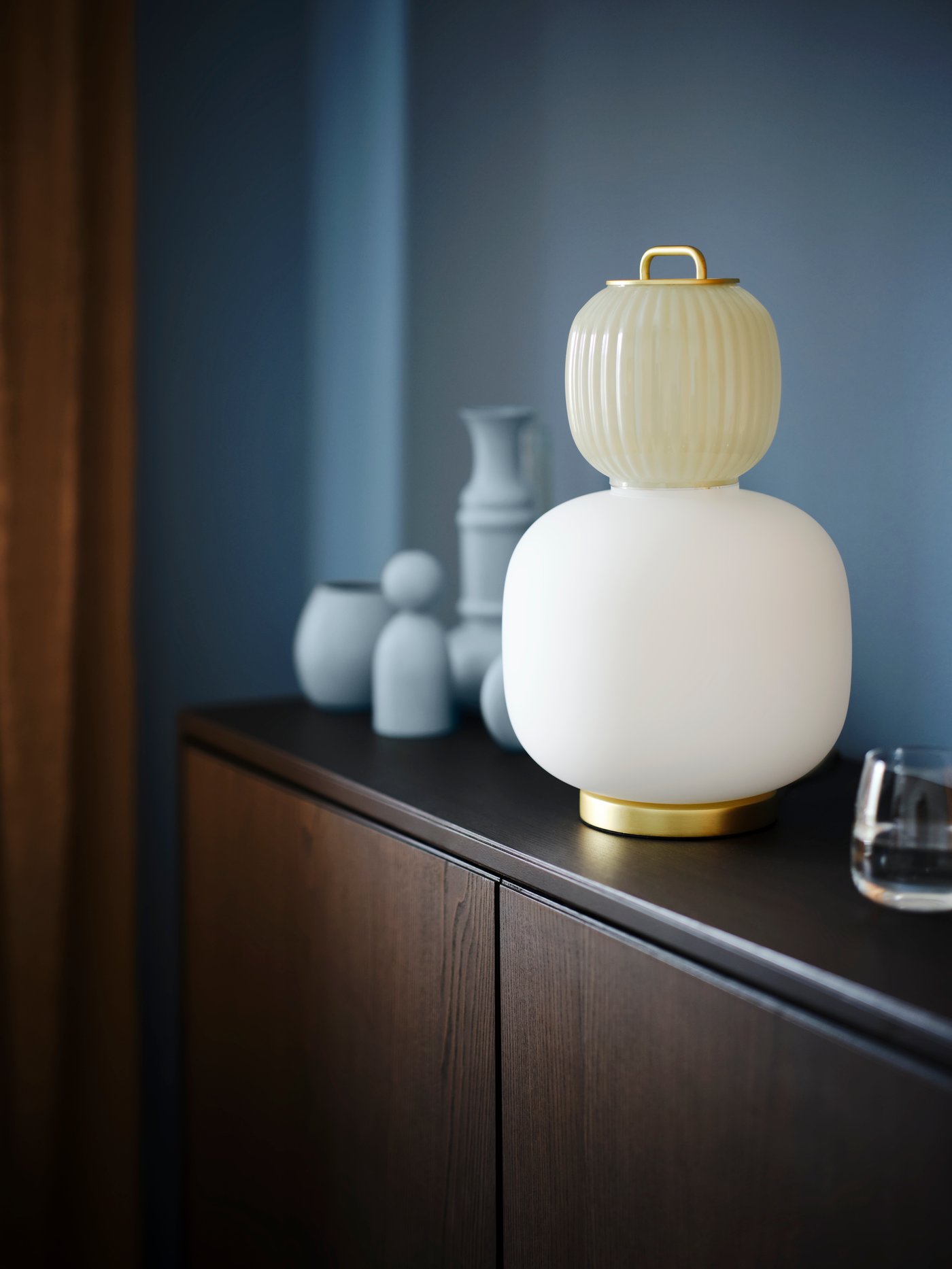 A brown REGISSÖR cabinet by a blue wall, with a white/light green glass PILBLIXT table lamp and a DYRGRIP glass on top.