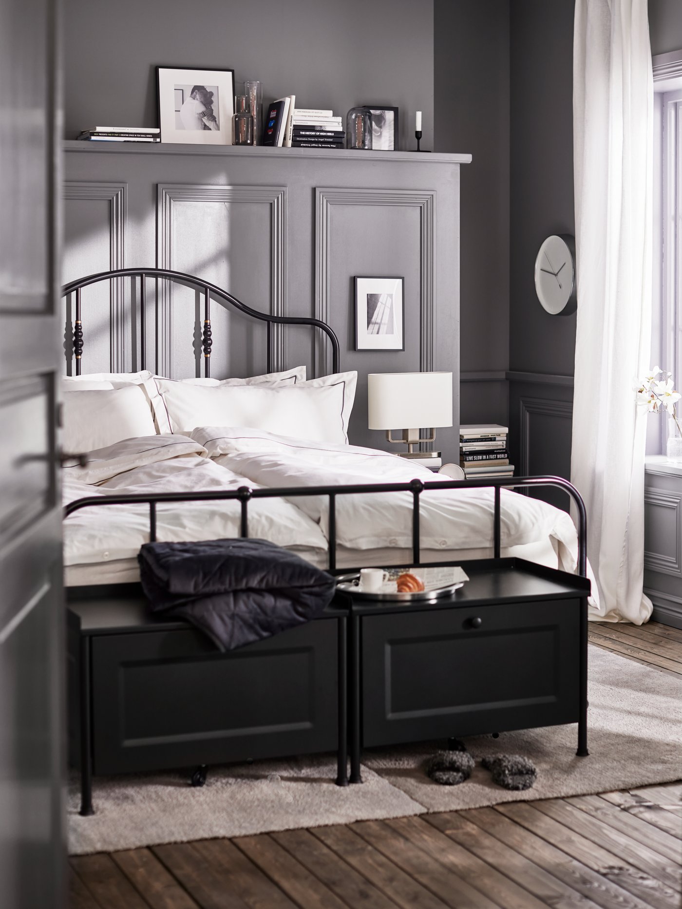Two black KORNSJÖ storage benches and a bed with a black bed frame made with white/dark grey bed linen on a light grey rug.