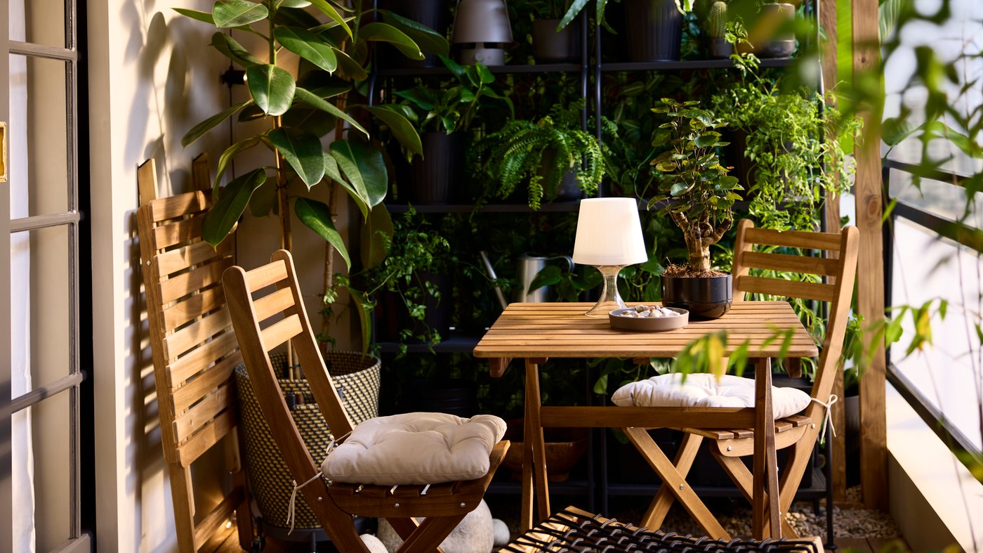 Een lichtbruin gebeitste ASKHOLMEN-klaptafel van acaciahout en twee klapstoelen staan op een balkon met planken en potplanten.