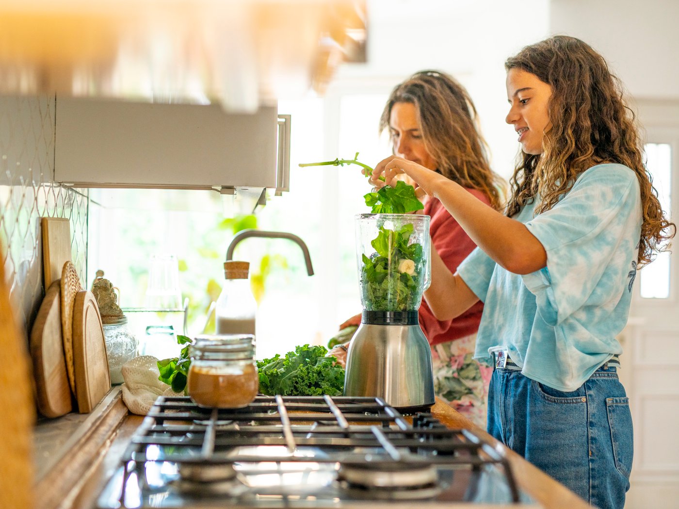 A mother and a teenage daughter chat in a kitchen, next to a blender full of greens.