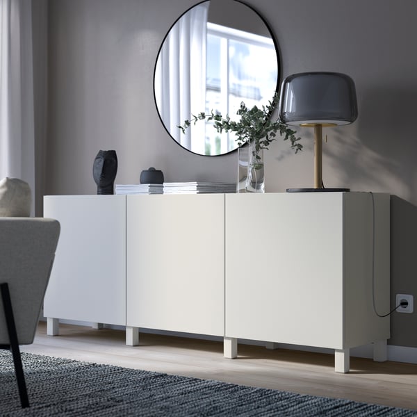 A white sideboard in front of a gray wall, with a large, round mirror above it. On the sideboard, on the right, is a table lamp.