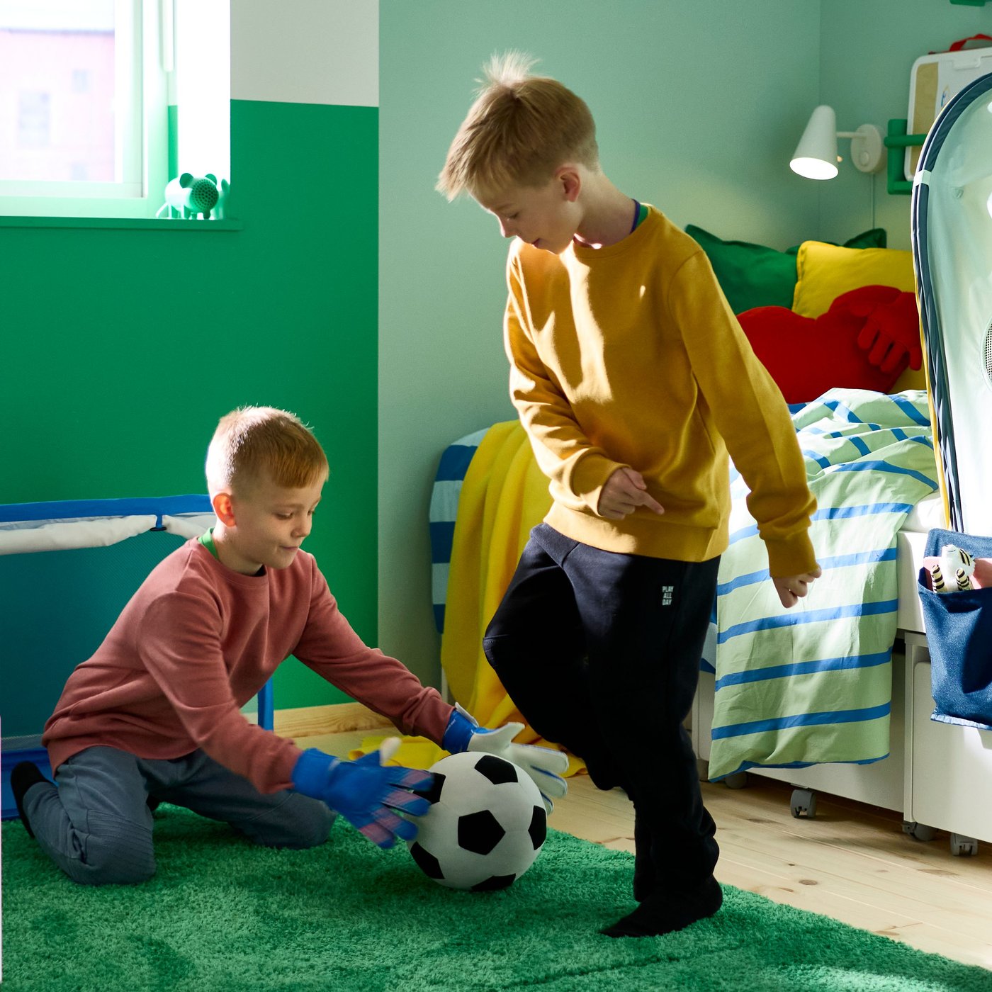 Un altoparlante Bluetooth portatile GREJSIMOJS verde, a forma di topolino, è appoggiato sul davanzale della finestra mentre due bambini giocano a calcio.