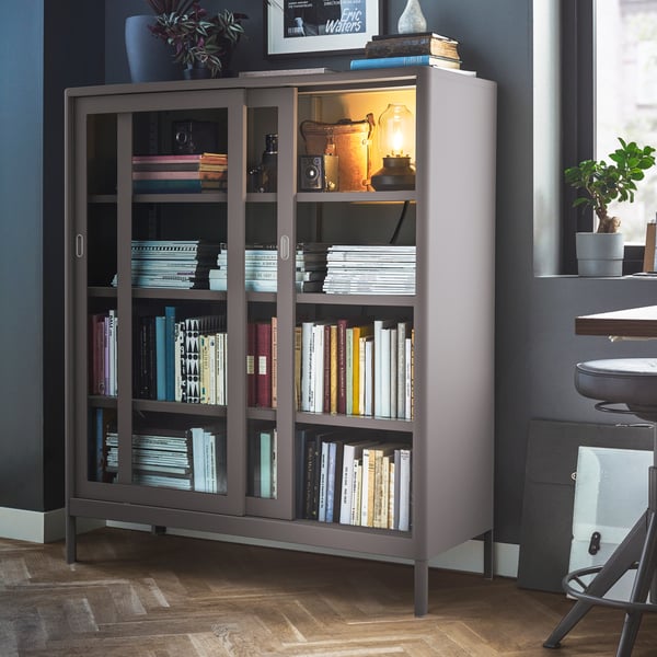 A parqueted room with a dark-grey IDÅSEN cabinet with sliding glass doors framed by art, decorations, plants and a window.