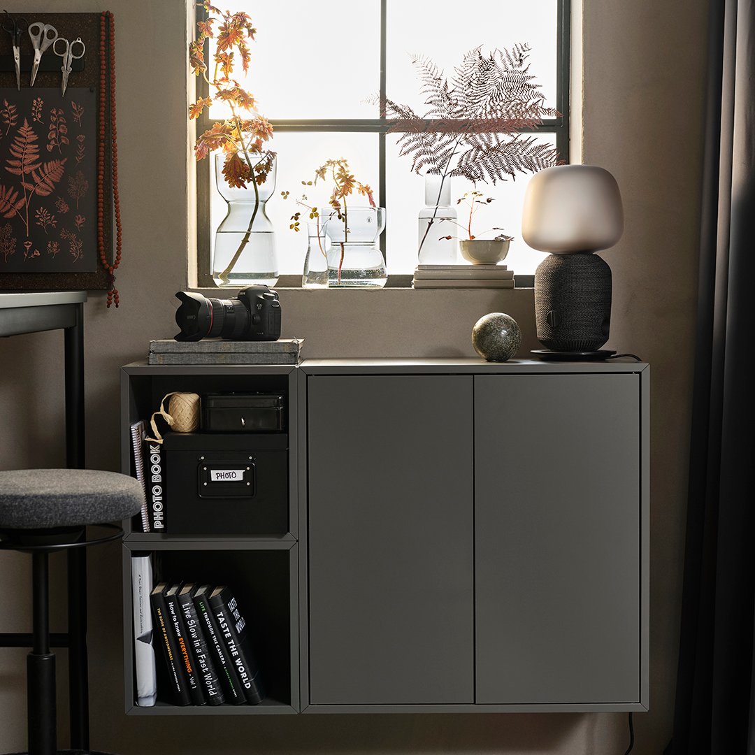 A dim room with a combination of dark-grey EKET cabinets mounted beneath a window. A WiFi speaker lamp is placed on top.