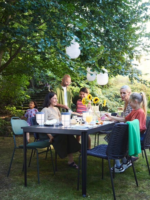 Dans un jardin, une famille s’apprête à manger, assise autour d’une table TEGELÖN installée sous un arbre.
